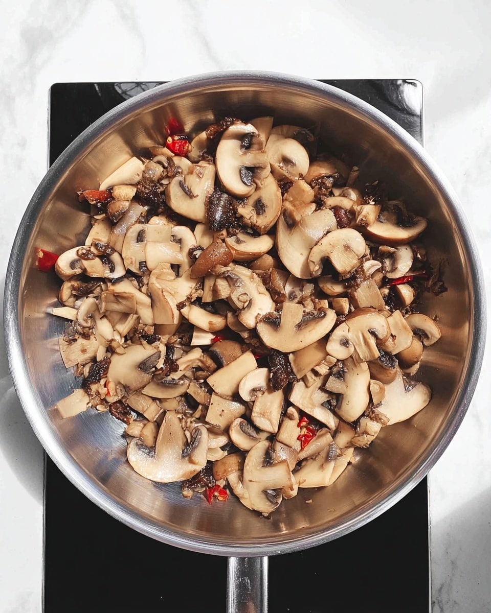 A shiny silver pan filled with cooked sliced mushrooms, showing light brown and beige tones with a glossy texture, mixed with darker pieces that look like dried mushrooms or spices and small red slices of chili scattered inside; the pan sits on a black stove surface against a white marbled texture background. photo taken with an iphone --ar 4:5 --v 7