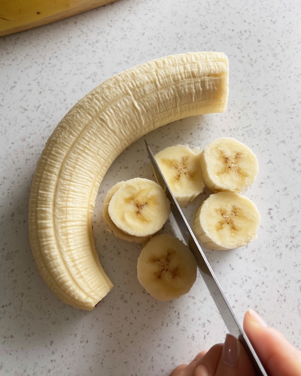 The image shows a peeled banana on a white marbled surface, with one half lying horizontally on top and the other half being sliced into round pieces. The sliced banana pieces are light yellow with soft, smooth texture and a few small brown spots in the center. A sharp knife is cutting through the banana, held by a woman's hand focusing on the banana near the bottom right of the image. The overall scene is bright and simple, emphasizing the fresh banana. Photo taken with an iphone --ar 4:5 --v 7