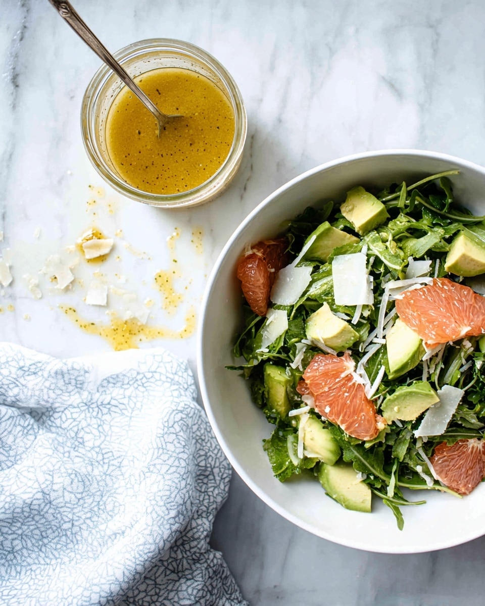 The image shows a white bowl filled with a fresh salad made of green leafy sprouts, slices of pale green avocado, and chunks of pink grapefruit, all topped with small pieces of white cheese shavings. Next to the bowl is a small clear glass jar filled with golden yellow salad dressing with a spoon inside. Around the jar, there are some spilled dressing drops and small bits of food on a white marbled surface. A folded light blue and white patterned cloth is placed near the jar, adding a soft texture to the scene. photo taken with an iphone --ar 4:5 --v 7