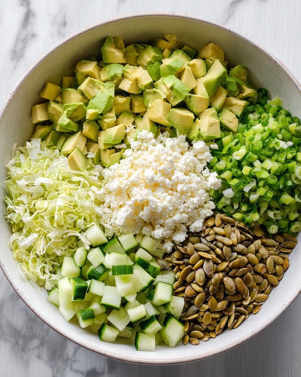 A large white bowl holds six separate layers of salad ingredients arranged side by side. Starting at the bottom left and moving clockwise, there is a layer of small chopped pale green cabbage pieces, next to it is a pile of light green chopped spring onions, followed by medium green diced cucumber with darker green skin, then light green to yellow diced avocado, a mound of creamy white crumbled cheese, and in the center a pile of shiny brown pumpkin seeds. The bowl is placed on a white marbled surface. Photo taken with an iphone --ar 4:5 --v 7