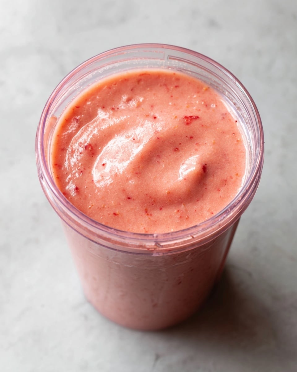 The image shows a clear blender cup filled with a smooth pink smoothie. The smoothie has a creamy texture with small specks of red and orange visible throughout. The cup is full almost to the top, and the surface of the smoothie is rounded with slight swirls. The cup is set on a white marbled surface. photo taken with an iphone --ar 4:5 --v 7