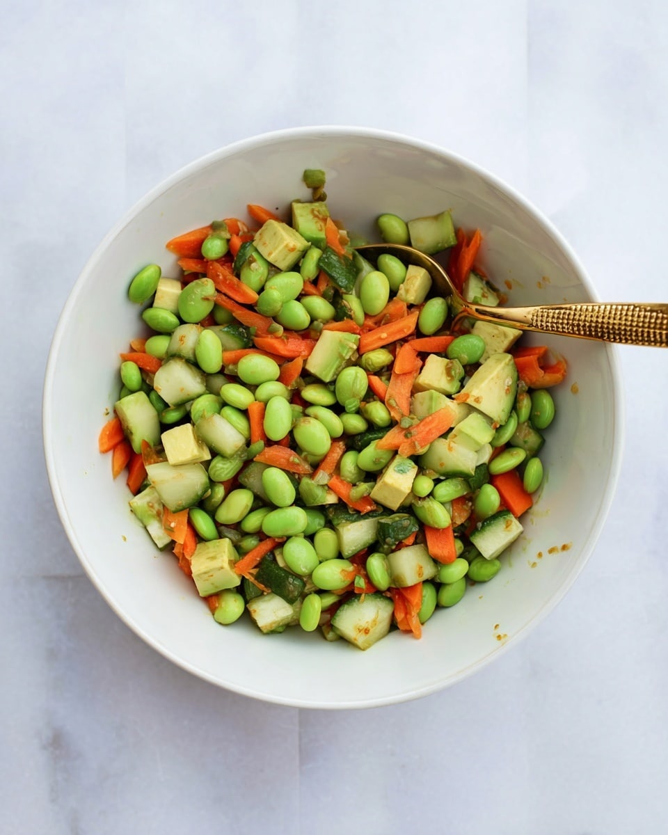 A white bowl filled with a colorful mix of vegetables, showing three main layers: bright green edamame beans scattered throughout, orange carrot pieces cut into small strips, and light green avocado chunks along with tiny cubes of cucumber. A golden spoon with a textured handle rests inside the bowl, partly submerged in the salad. The bowl sits on a white marbled surface, giving a fresh and clean look to the image. Photo taken with an iphone --ar 4:5 --v 7