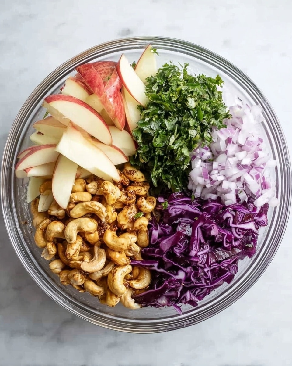 A clear glass bowl containing five distinct layers arranged in separate sections: thin slices of red apple with pale cream flesh at the top left, finely chopped light purple onion pieces to the top right, a pile of chopped fresh green herbs on the right side, a layer of chopped deep purple cabbage at the bottom left, and a mound of roasted cashew nuts with a golden brown, slightly oily texture in the center. The bowl is placed on a white marbled surface. Photo taken with an iphone --ar 4:5 --v 7