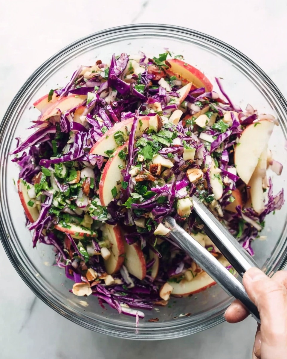 A clear glass bowl filled with a colorful salad showing three main layers: thin strips of purple cabbage giving a vibrant deep purple color, thin apple slices with red skin adding a light cream and red contrast, and chopped green herbs scattered on top for fresh green touches. There are also small chunks of light brown nuts and tiny pieces of finely chopped white onion mixed throughout. A woman's hand using silver tongs is lifting a portion of the salad, capturing the mix of all the ingredients closely against a white marbled surface background photo taken with an iphone --ar 4:5 --v 7