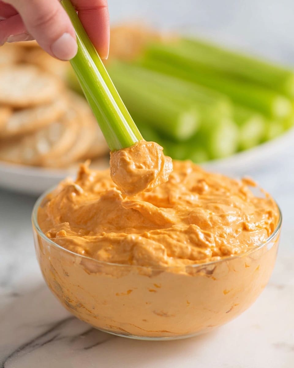 A clear round glass bowl filled with a thick, creamy orange dip with visible small chunks, sitting on a white marbled surface. Above the bowl, a bright green celery stick is held by a woman's hand, dipped into the orange spread, showing a generous amount clinging to the celery's notch. In the blurred background, there is a white plate with more celery sticks, and some light brown crackers are also faintly visible. The scene is brightly lit and focused on the texture of the dip and celery. photo taken with an iphone --ar 4:5 --v 7