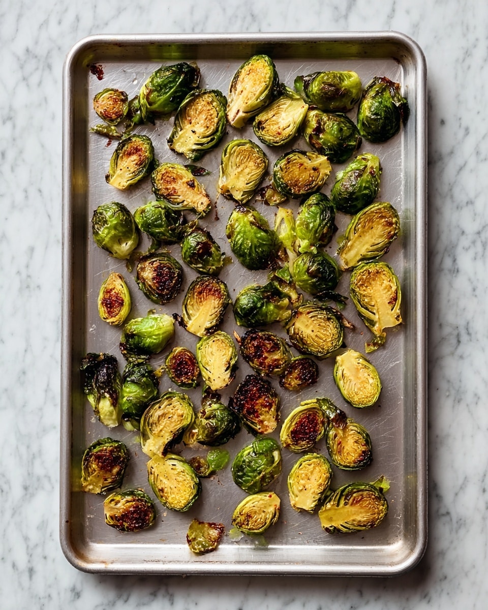 A silver baking tray filled with many halved Brussels sprouts, each with a green outer layer and a browned, slightly crispy yellow-green inside. The Brussels sprouts are spread out unevenly across the tray, some showing darker char marks on their cut sides while others have a mix of green and brown tones from roasting. The tray rests on a surface with a white marbled texture. photo taken with an iphone --ar 4:5 --v 7