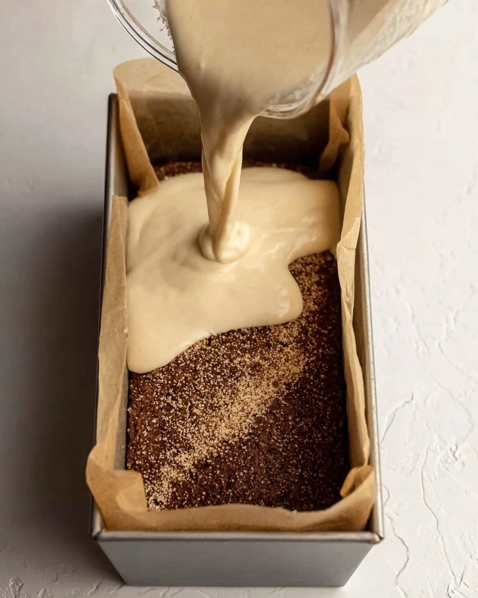 A loaf pan lined with parchment paper is filled with a dark brown crumb layer at the bottom, which looks coarse and even. A light beige batter with smooth, creamy texture is being poured from a glass measuring cup in the top right corner, flowing softly over the crumb layer. The background and surface show a white marbled texture. photo taken with an iphone --ar 4:5 --v 7