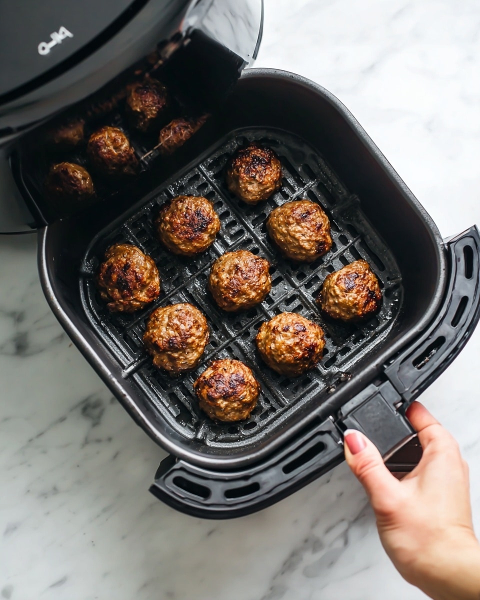 The image shows a black air fryer basket with nine small, round, browned meatballs inside. The meatballs are cooked with a slightly crispy and charred outside texture. The basket has a grid pattern at the bottom for air flow, and it is being pulled out by a woman's hand on the right side. The background is a white marbled surface. The air fryer lid is partially lifted at the top left corner, reflecting a faint image of the meatballs. Photo taken with an iphone --ar 4:5 --v 7