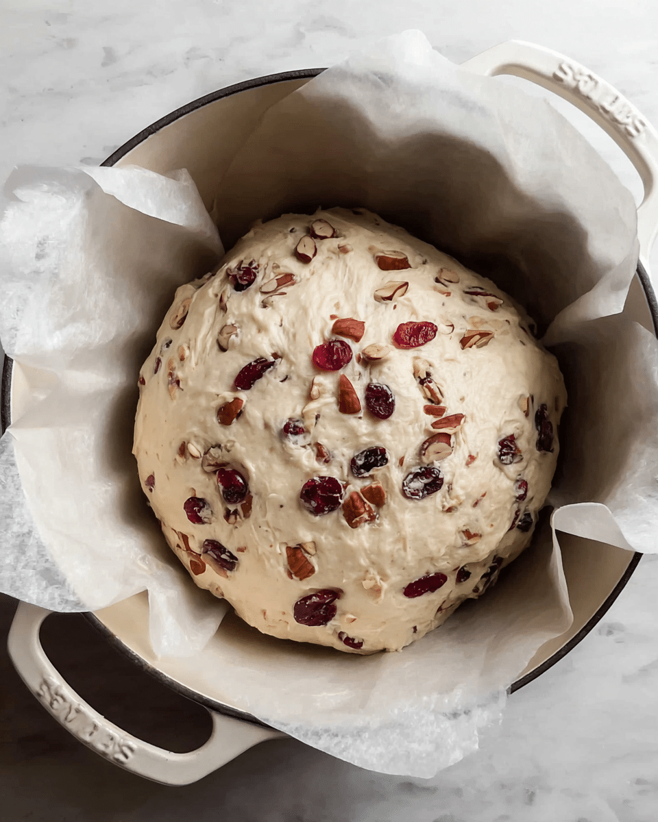 A round dough ball mixed with dried cranberries and nuts is resting inside white parchment paper. The dough is pale and soft with embedded dark red cranberries and light brown nuts scattered within and on top. The parchment paper cradles the dough with gently folded edges rising above it, sitting inside a white cast iron pot with visible handles. The pot is placed on a white marbled surface. photo taken with an iphone --ar 4:5 --v 7