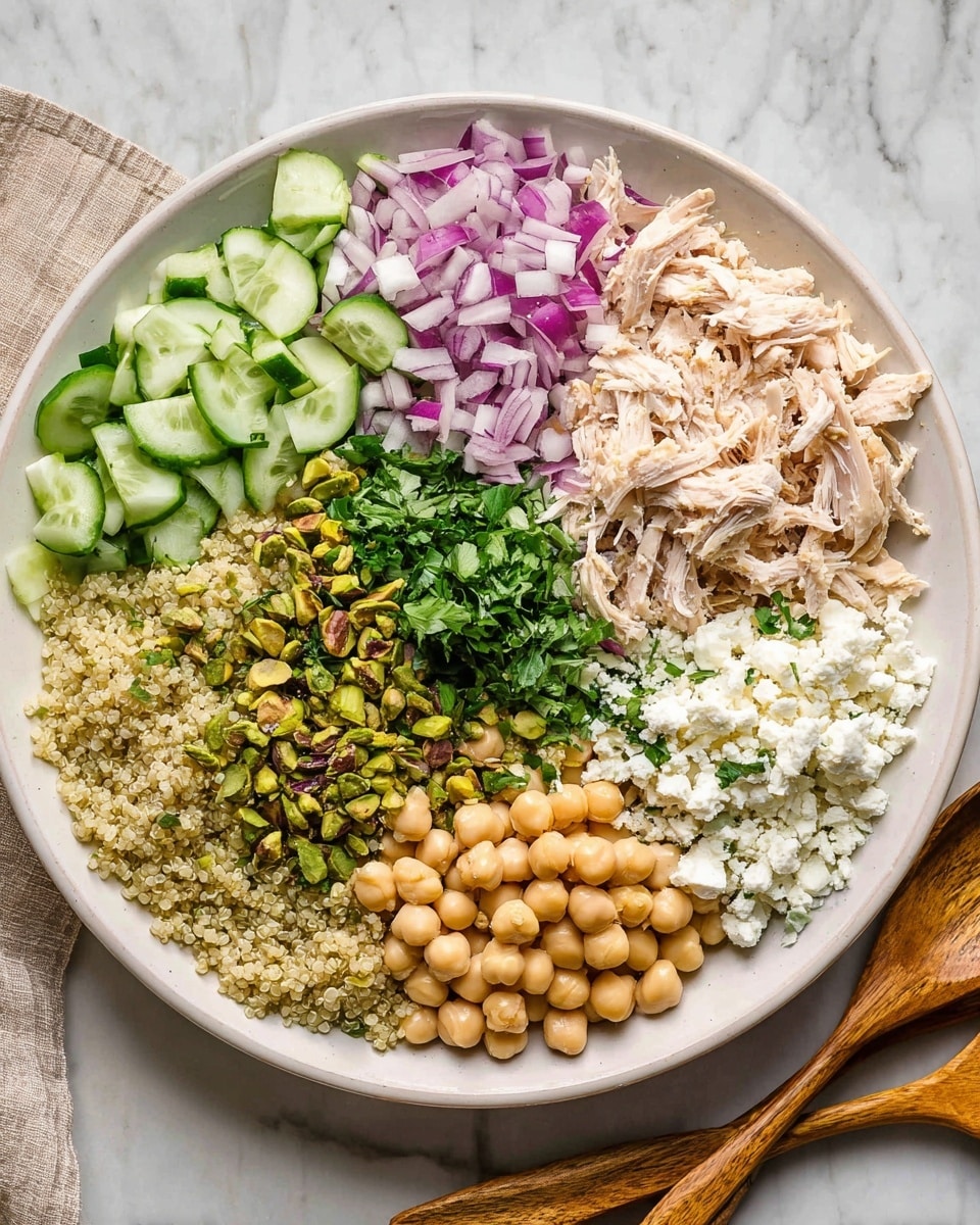 A white plate with seven layers arranged in a circle: at the top right shredded light pink chicken, next to it finely diced purple and white onions, below that a pile of green pistachios, followed by crumbled white cheese at the bottom right, then round beige chickpeas at the bottom center, cooked light yellow quinoa to the bottom left, thinly sliced light and dark green cucumber at the top left, and finely chopped fresh green herbs in the center. The plate sits on a white marbled surface, with two wooden salad utensils lying nearby. Photo taken with an iphone --ar 4:5 --v 7