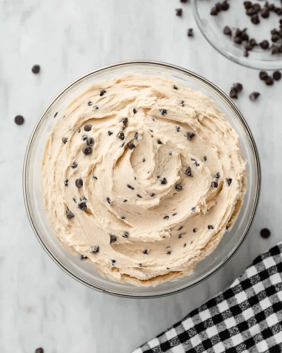 A clear glass bowl sits on a white marbled surface, filled with a single thick layer of creamy, light beige dough mixed with small dark chocolate chips evenly spread throughout. A black and white checked cloth is partly visible in the top right corner, with scattered chocolate chips lying around the bowl. The dough texture looks soft and smooth, with gentle swirls showing where it has been stirred. photo taken with an iphone --ar 4:5 --v 7