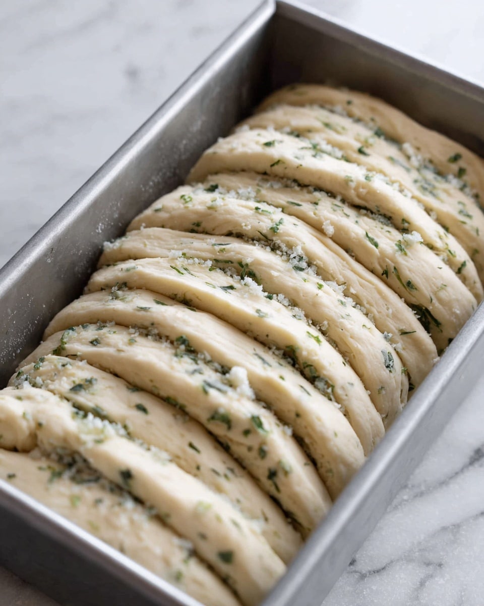 The image shows a silver baking pan filled with multiple layers of folded dough pieces stacked next to each other. Each layer is light beige in color with flecks of green herbs and white flakes spread evenly between them, giving a soft and fluffy texture. The dough layers curve gently and are arranged in a neat row, showing the texture of herbs and salt inside. The baking pan rests on a surface with a white marbled texture. Photo taken with an iphone --ar 4:5 --v 7
