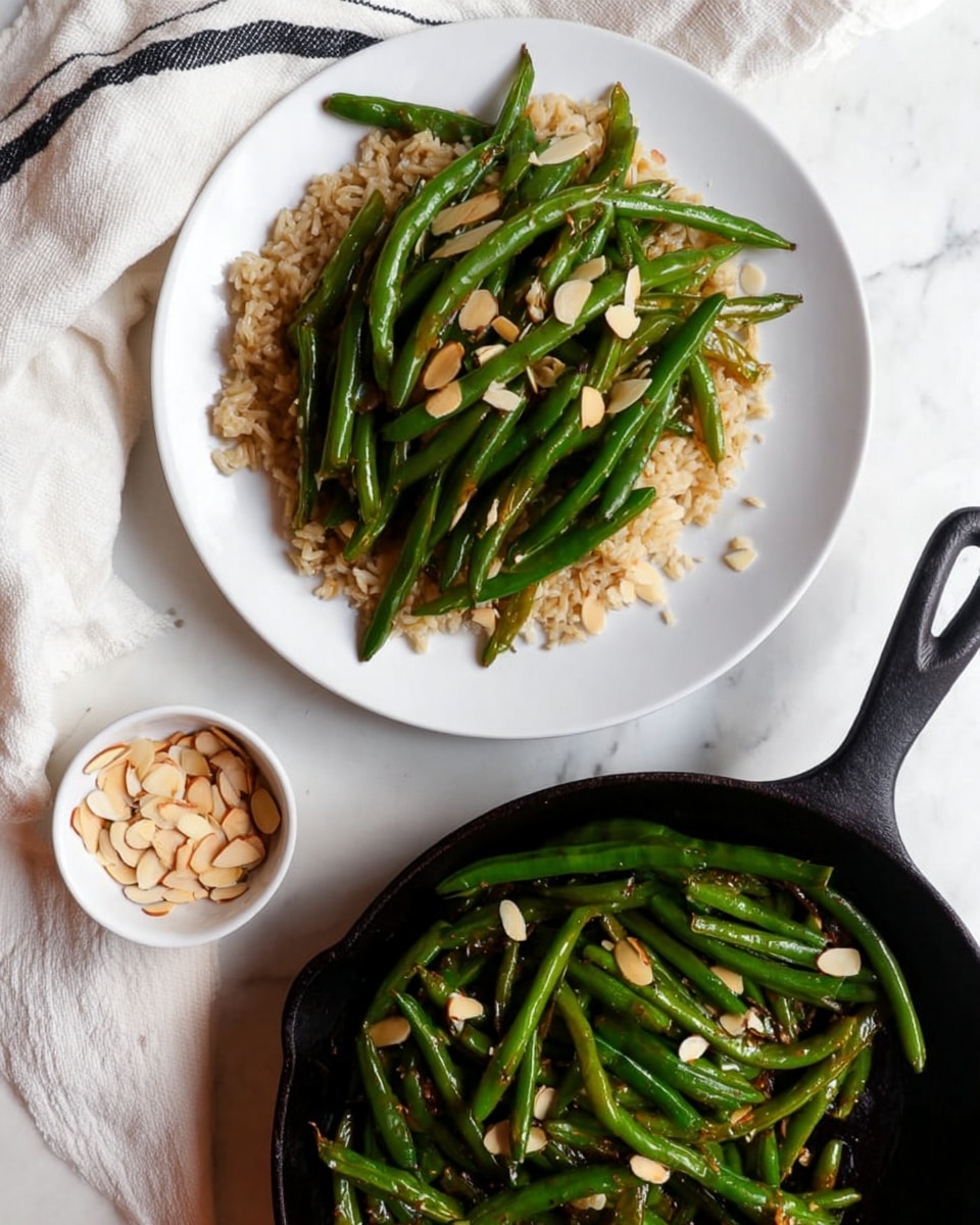 A white round plate holds a base layer of light brown cooked rice, topped with a layer of bright green cooked green beans mixed with small slivers of white almonds, some green beans have a light shine from a sauce. To the side on a white marbled surface is a small white bowl filled with more almond slivers. Below the plate is a black cast iron skillet containing more green beans cooked in the same sauce and garnished with almond slivers. The background includes a white cloth with black stripes on the edge of the skillet. Photo taken with an iphone --ar 4:5 --v 7