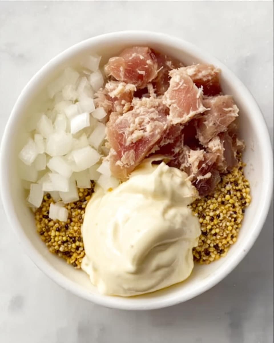 A white bowl holds four main visible layers arranged separately: on the top right, there is a light brown chunk of tuna; on the top left, finely chopped white onions; below the tuna, a dollop of coarse grain mustard with visible mustard seeds in yellow and dark brown shades; and below the mustard, a large spoonful of creamy white mayonnaise. All ingredients have distinct textures and colors that contrast clearly against the white bowl and a white marbled surface in the background. Photo taken with an iphone --ar 4:5 --v 7