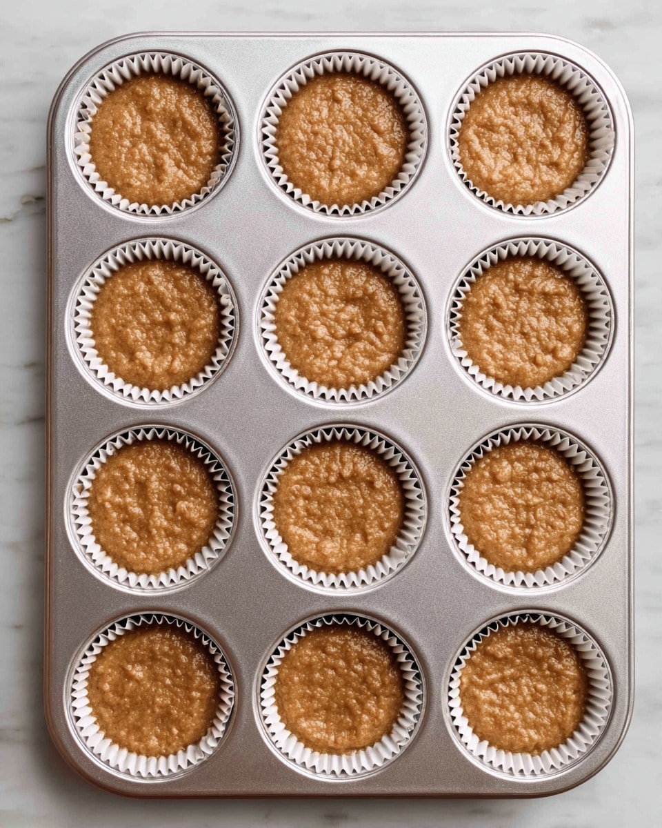 A silver cupcake tray with twelve round cups holds twelve paper liners filled with light brown batter, each liner showing ridged edges and the batter slightly rough on top with a few small lumps, evenly distributed and ready to bake, all placed on a white marbled surface photo taken with an iphone --ar 4:5 --v 7