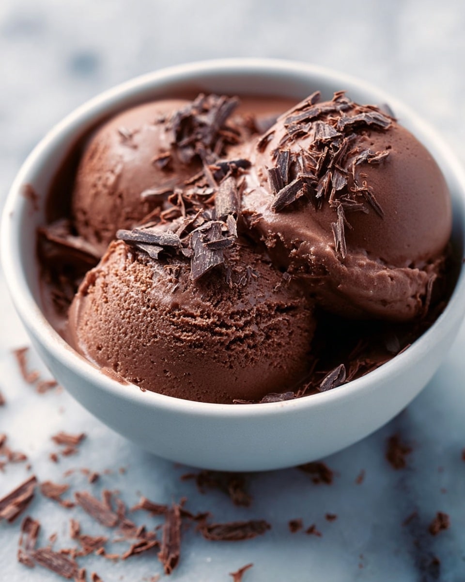 A close-up view of a white bowl filled with two smooth, round scoops of rich brown chocolate mousse, topped with small flakes of dark chocolate scattered evenly on the surface and around the bowl. The mousse looks creamy and dense, with a shiny texture that reflects light softly. The bowl is placed on a white marbled surface, and some chocolate flakes are scattered outside the bowl, adding a natural and slightly messy touch. photo taken with an iphone --ar 4:5 --v 7