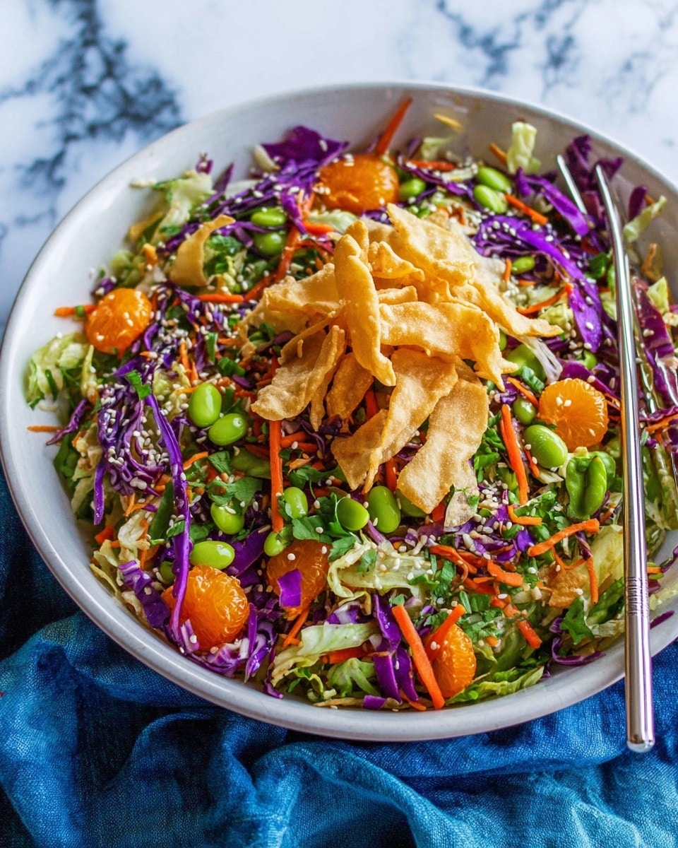 The dish is a colorful salad served in a white bowl sitting on a white marbled surface with a blue cloth partially underneath. The bottom layer of the salad is made up of shredded green lettuce and purple cabbage, mixed with thin orange carrot strips. Scattered throughout are bright green edamame beans and pieces of leafy green herbs. There are also small orange segments spread across the salad. On top of all these layers is a pile of light tan crispy fried strips, adding texture. White sesame seeds are sprinkled over the salad, and a pair of metal chopsticks rests on the white bowl. Photo taken with an iphone --ar 4:5 --v 7