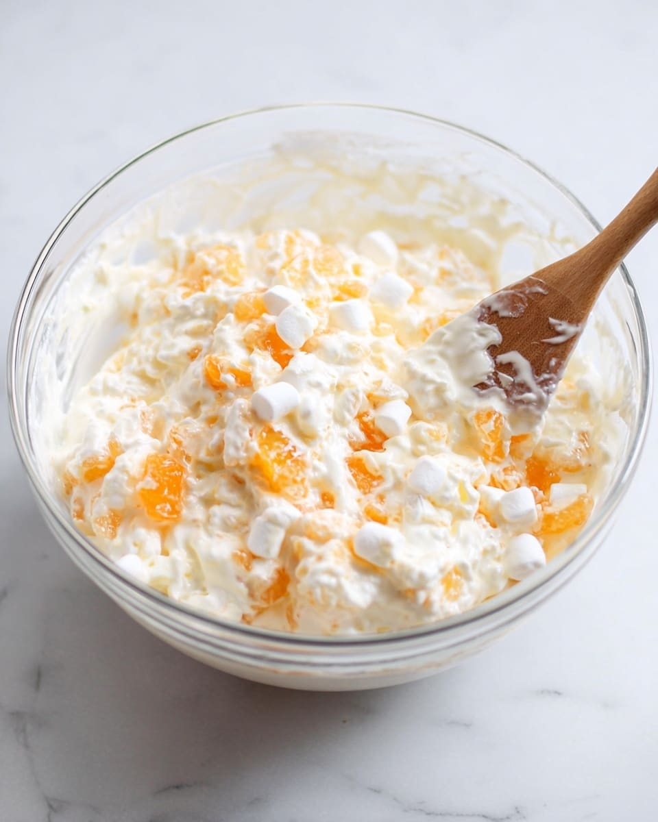 A clear glass bowl filled with a creamy mixture containing white small marshmallows, soft white cottage cheese, and bright orange small fruit pieces, likely mandarin, all evenly mixed together. A spoon with a dark handle is partly dipped in the mixture, resting against the bowl's edge. The bowl is placed on a white marbled texture surface. photo taken with an iphone --ar 4:5 --v 7