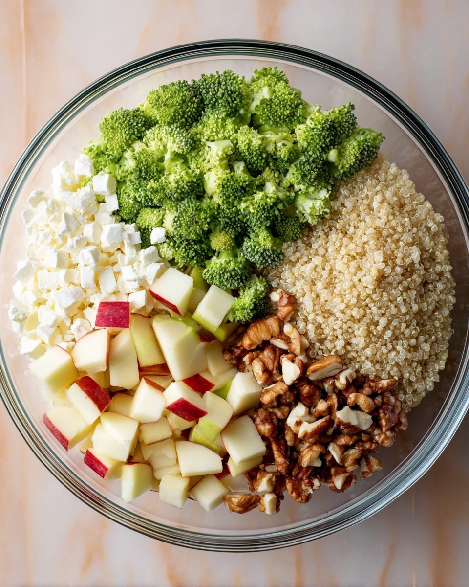 A clear glass bowl holds five distinct layers arranged in separate sections: at the top, bright green chopped broccoli florets with a fresh, textured look; next to it, a white crumbly layer of cheese with a soft texture; below the cheese, pale yellow and red small apple cubes with smooth surfaces; moving left, a mix of chopped brown nuts showing varied sizes and rough textures; and finally, cooked light beige quinoa grains with a slightly fluffy and grainy texture. The bowl sits on a white marbled surface. photo taken with an iphone --ar 4:5 --v 7