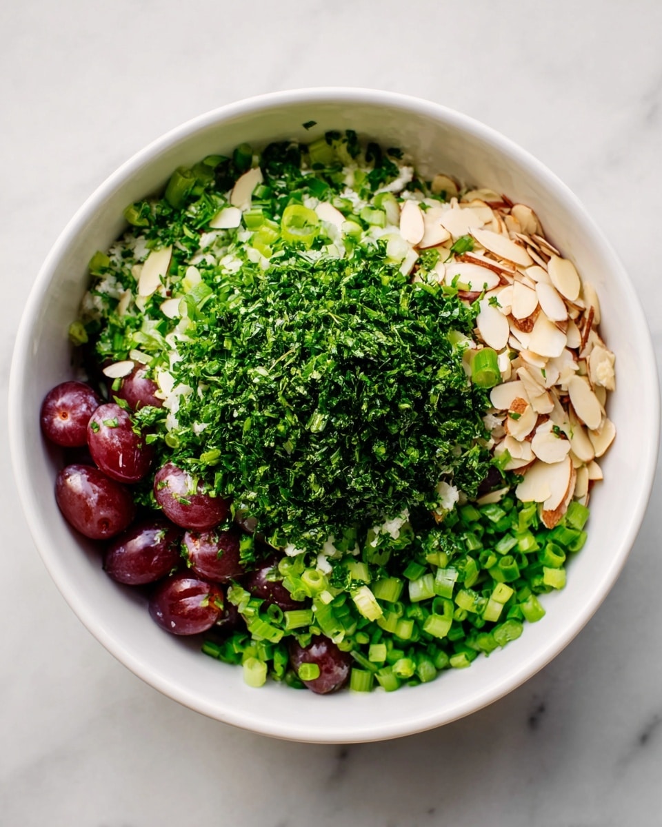 A white bowl filled with multiple layers of fresh ingredients on a white marbled surface. The bottom layer consists of sliced red grapes visible at the edges. On top of the grapes, there is a layer of pale sliced almonds spread evenly. Above the almonds, there is a thick layer of finely chopped green onions covering most of the bowl. The topmost layer is a large mound of finely chopped dark green herbs placed roughly in the center. The textures show a mix of soft grapes, crunchy almonds, and fresh green herbs. Photo taken with an iphone --ar 4:5 --v 7