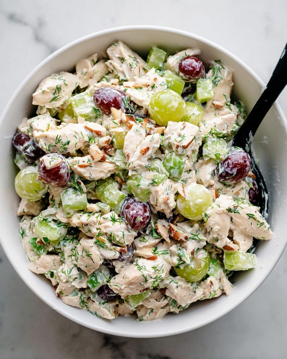 A white bowl filled with a mixed salad showing layers of creamy, pale beige chicken pieces, bright green celery chunks, deep red sliced grapes, and light brown sliced almonds, all coated in a creamy white dressing with green herb specks evenly spread throughout. A black spoon is partially inside the bowl on the right side. The bowl is placed on a white marbled surface. photo taken with an iphone --ar 4:5 --v 7