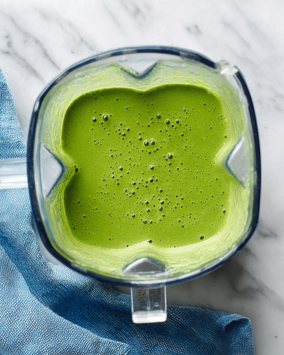 A clear blender container filled with a smooth, bright green sauce or smoothie with a slightly frothy top and small bubbles across the surface, placed on a white marbled surface with a folded blue cloth nearby photo taken with an iphone --ar 4:5 --v 7