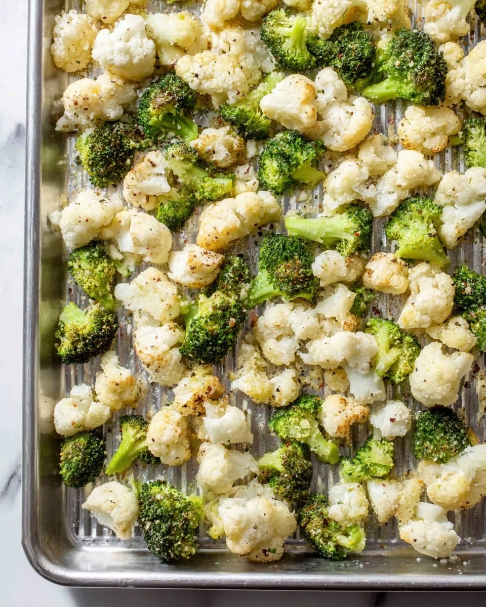 The image shows a baking tray filled with many small pieces of cauliflower and broccoli, evenly spread in a single layer. The cauliflower pieces are off-white with a slightly crumbly texture, while the broccoli pieces are green with a bumpy, firm look. Both vegetables have a light sprinkling of seasoning that adds small specks of color. The tray itself is metallic with visible ridges, placed on a white marbled surface. Photo taken with an iphone --ar 4:5 --v 7