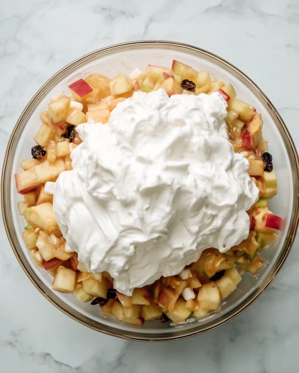 A clear glass bowl sits on a white marbled surface, filled with small chopped pieces of apples and other fruits mixed with a few raisins and small marshmallows, creating a mix of light yellow, red, and beige colors with some dark spots. On top of this fruit mix, there is a large dollop of thick white whipped cream that contrasts with the colorful fruit beneath. The bowl shows layers of textured fruit pieces under the smooth, fluffy cream layer. photo taken with an iphone --ar 4:5 --v 7