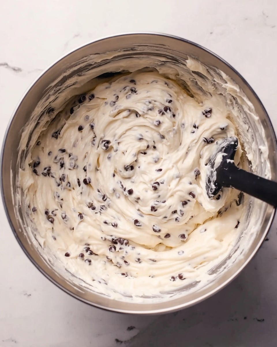 A metal mixing bowl filled with creamy white batter that is thick and smooth, dotted evenly with many small dark chocolate chips throughout. The batter has soft swirls and texture from stirring, and a black spatula is partially visible inside the bowl resting on the batter. The bowl is placed on a white marbled surface. Photo taken with an iphone --ar 4:5 --v 7