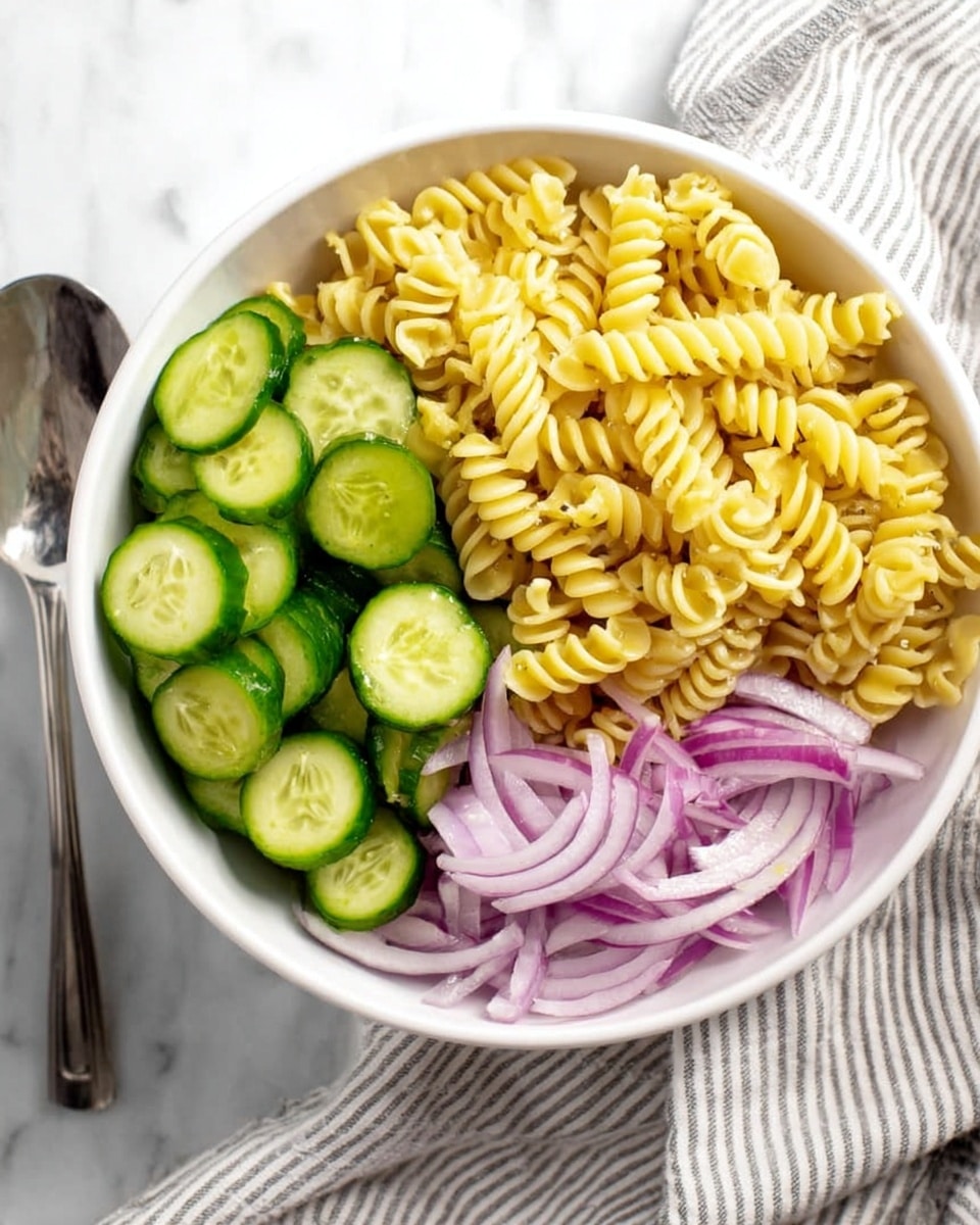 A white bowl filled with three clear sections: one section has light yellow twisted pasta spirals, another section holds bright green round cucumber slices, and the third section contains thin, slightly translucent purple onion slices with white edges, all arranged neatly without mixing. The bowl is placed on a white marbled surface with a striped cloth and shiny silver spoon nearby, photo taken with an iphone --ar 4:5 --v 7