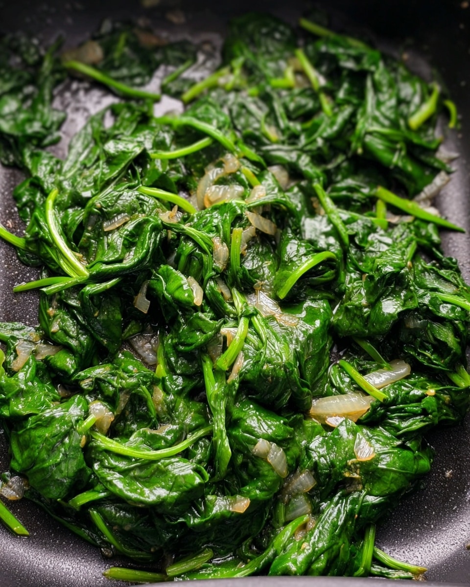 A close-up view of cooked spinach mixed with small pieces of soft, translucent onions in a dark pan. The spinach leaves are vibrant green with some wilted, shiny, and slightly wrinkled textures. The stems of spinach are still attached and scattered evenly among the tender onion bits. The pan has a smooth, dark surface with a slight oil sheen, highlighting the cooked vegetables. The image shows a simple, single layer of the spinach and onions filling the pan. photo taken with an iphone --ar 4:5 --v 7