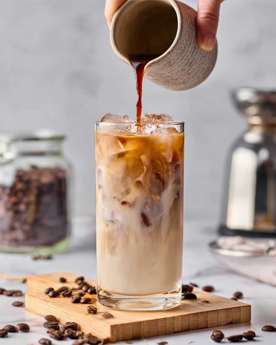 A tall clear glass filled with iced milk, showing white and light brown layers with ice cubes inside. A woman's hand pours dark brown coffee from a small textured light gray cup into the top of the glass, creating a swirling mix of colors. The glass sits on a light wooden block with scattered dark brown coffee beans around it, all placed on a white marbled surface. In the background, a clear glass container with coffee beans and a silver coffee maker are slightly blurred. photo taken with an iphone --ar 4:5 --v 7