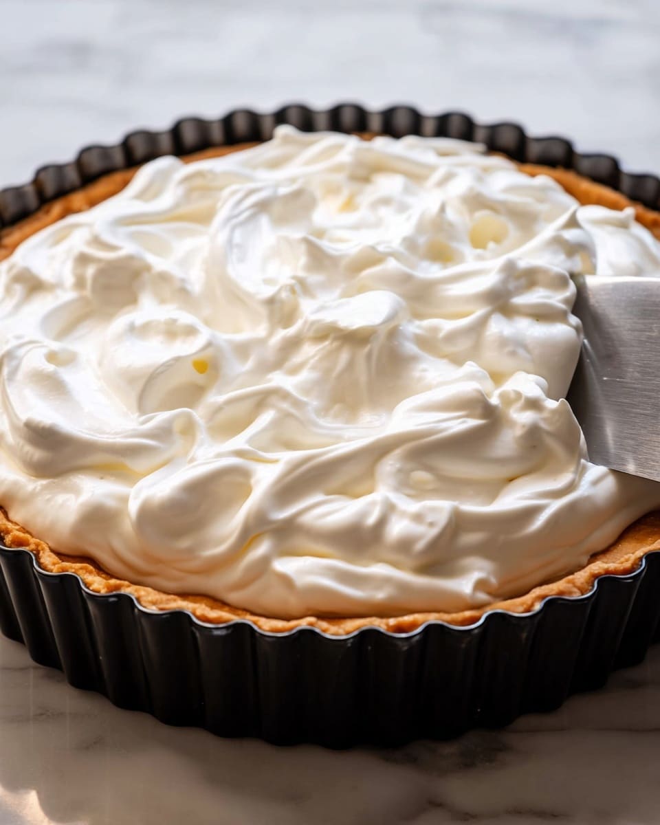 The image shows a pie with a single visible layer of thick, white whipped cream topping that is spread thickly with soft waves and peaks on top. The pie crust below is golden brown and fits snugly into a black fluted tart pan with wavy edges. A shiny silver spatula is gently smoothing the cream on the right side. The entire pie sits on a white marbled surface in soft natural light. photo taken with an iphone --ar 4:5 --v 7