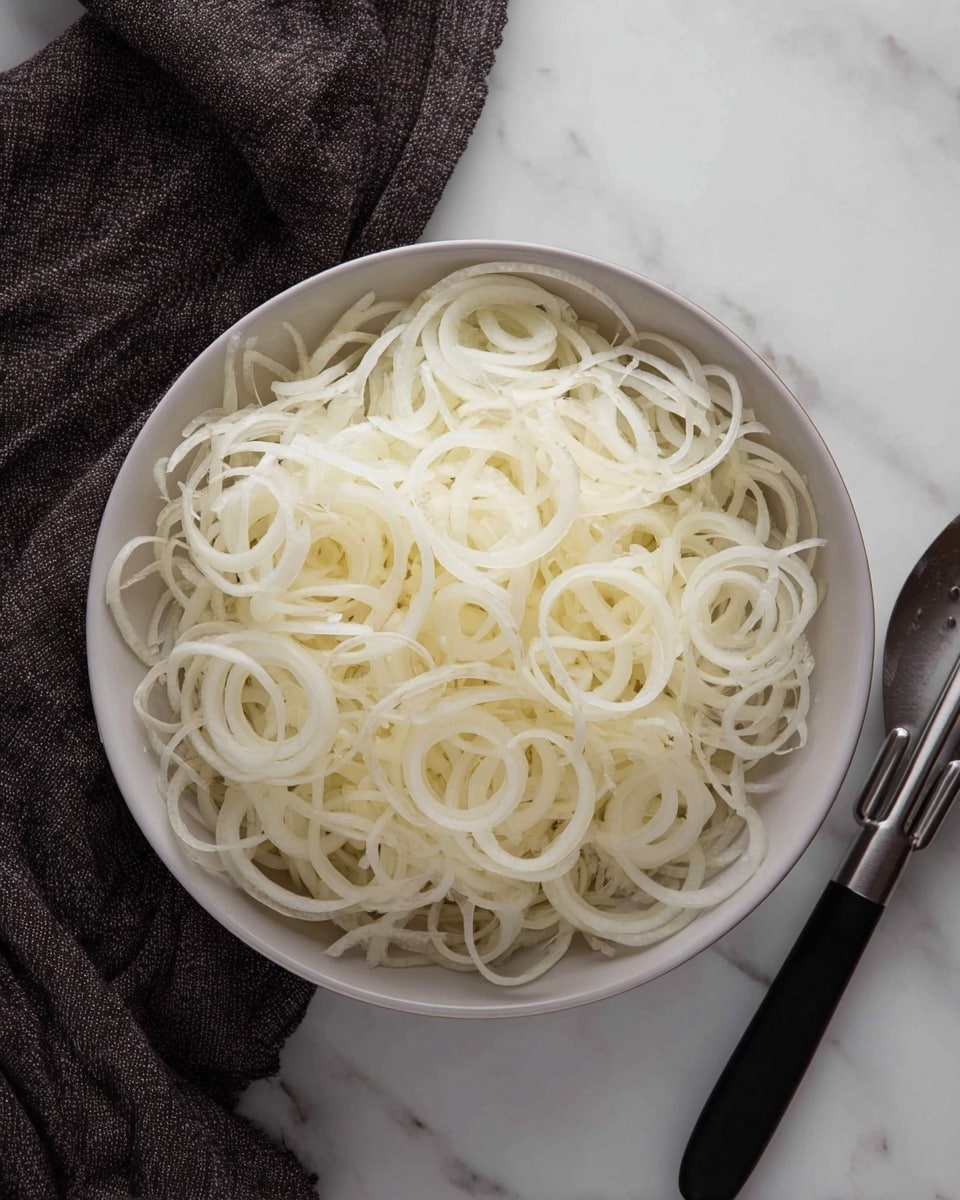 A round white bowl filled with many thin, white onion rings piled loosely inside. The thin layers of onion rings create a curly, spiral texture all over, and the bowl sits on a white marbled surface. Near the bowl, there is a silver tong with a black silicone tip resting on the surface, and a dark grey cloth is partially visible near the top edge of the image. photo taken with an iphone --ar 4:5 --v 7