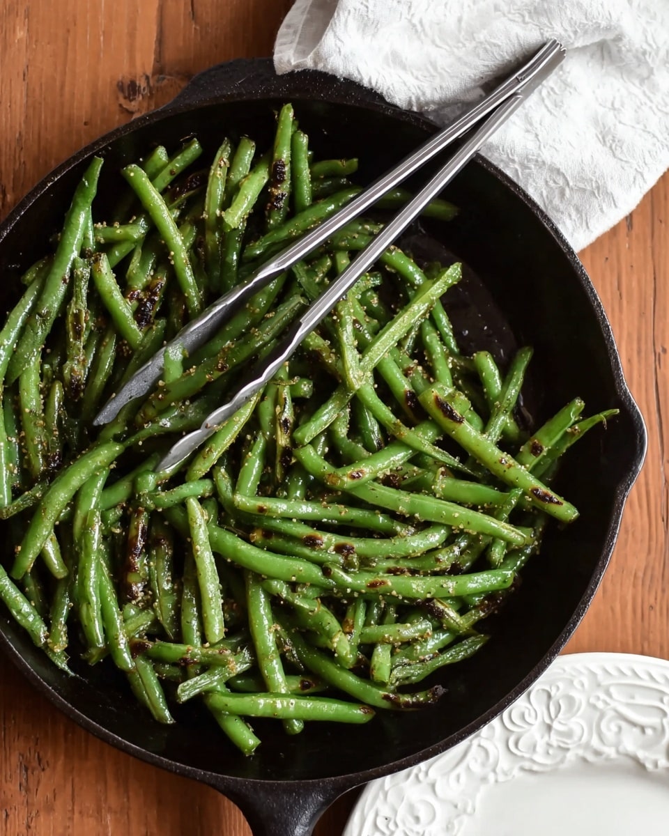 A black cast iron pan is filled with bright green cooked green beans that have small brown grill marks and some coarse seasoning visible on them. A pair of silver tongs rests diagonally inside the pan on top of the beans. The pan is placed on a wooden surface next to a white cloth napkin and a white plate with an embossed floral edge, all set on a white marbled surface. Photo taken with an iphone --ar 4:5 --v 7