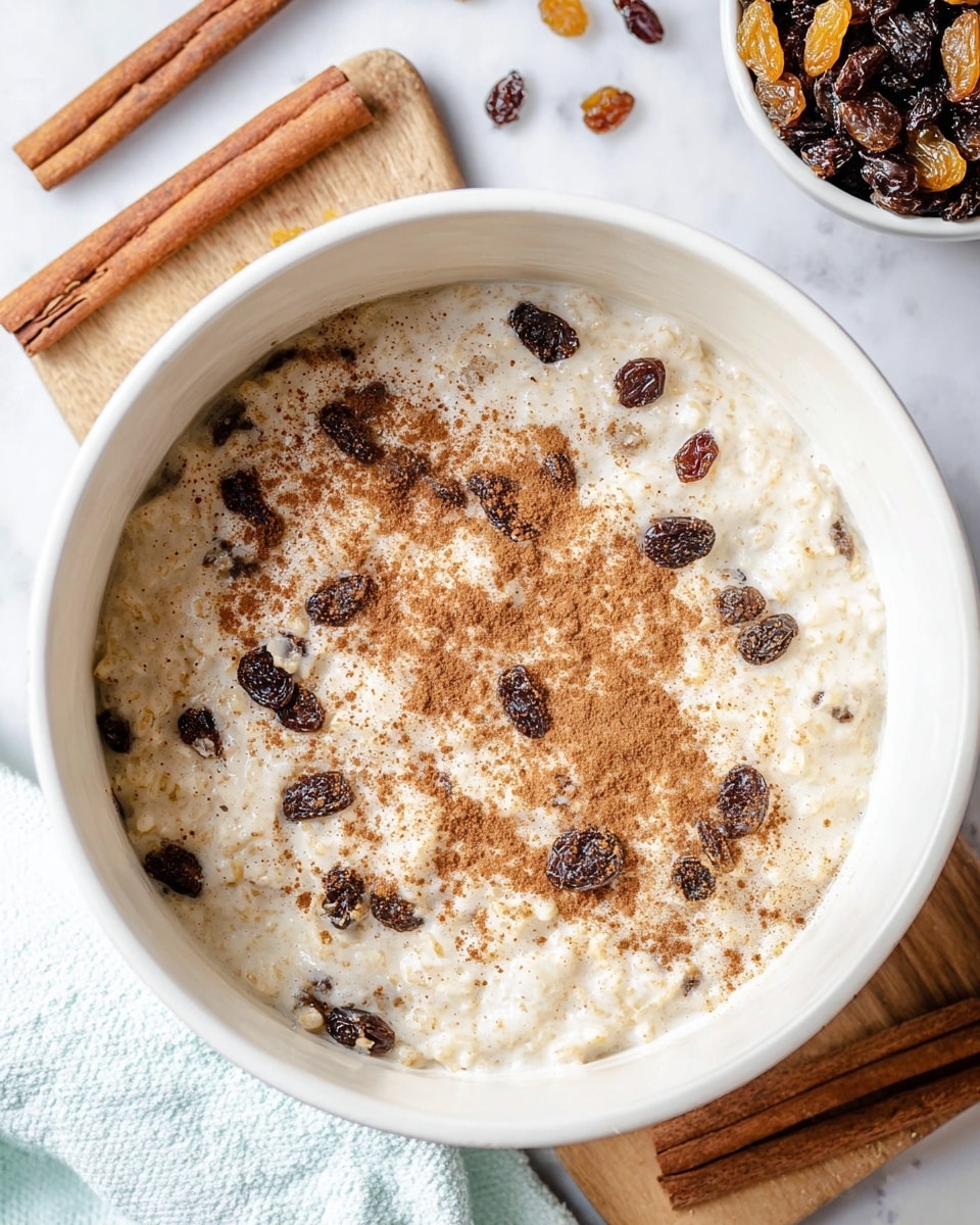 The image shows a white pot filled with creamy rice pudding mixed with dark brown raisins. The surface of the pudding is sprinkled lightly with brown cinnamon powder in small patches. The pot sits on a white marbled surface with a few cinnamon sticks and scattered raisins nearby. Part of a white cloth with a light blue edge is seen at the bottom left corner. There is also a white bowl filled with dark and golden raisins placed near the top right corner of the pot photo taken with an iphone --ar 4:5 --v 7