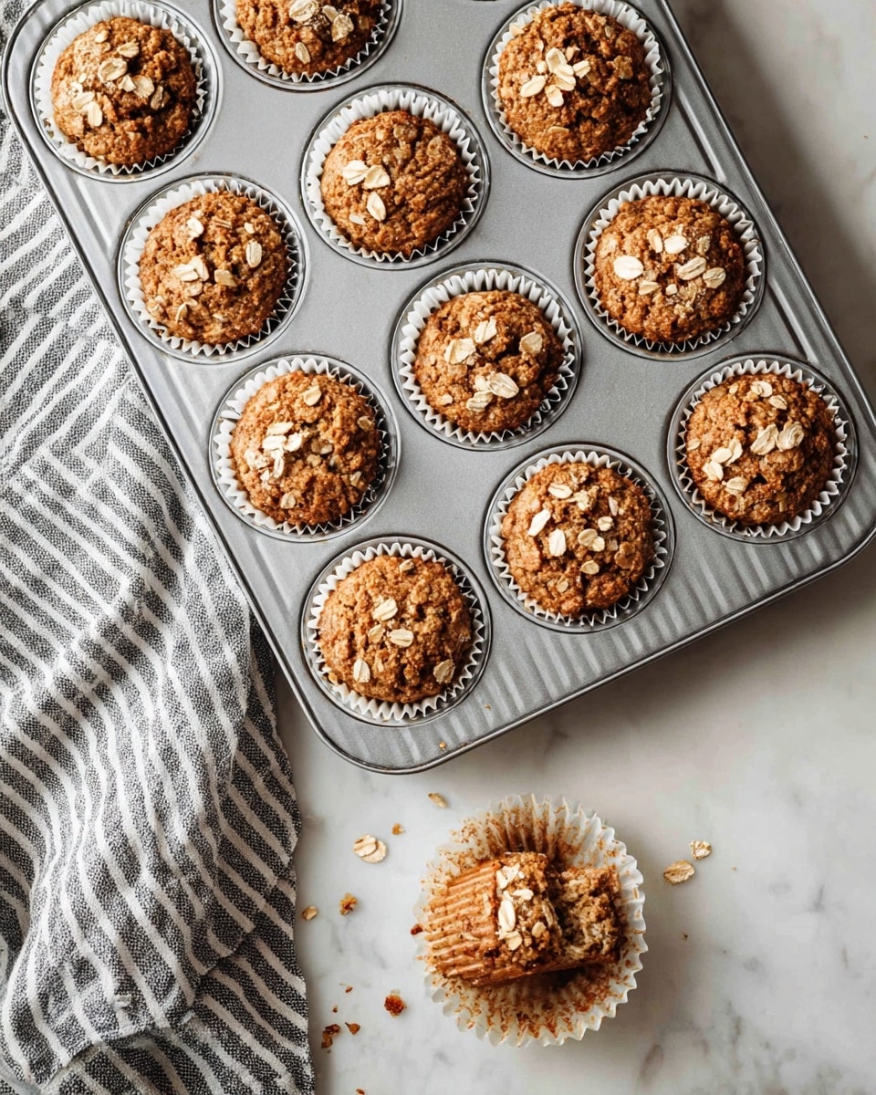 The image shows a metal muffin tray with ten golden brown muffins, each topped with large oats. The muffins are in white paper liners and have a textured, slightly cracked surface with visible pieces of oats on top. One muffin is out of the tray and placed below on a white marbled surface, slightly broken apart to show the soft, moist inside with small bits of ingredients visible. To the left side of the tray, there is a gray and white striped cloth on the white marbled surface. The whole setup rests on a white marbled countertop, and the lighting is soft, highlighting the warm tones of the muffins. photo taken with an iphone --ar 4:5 --v 7
