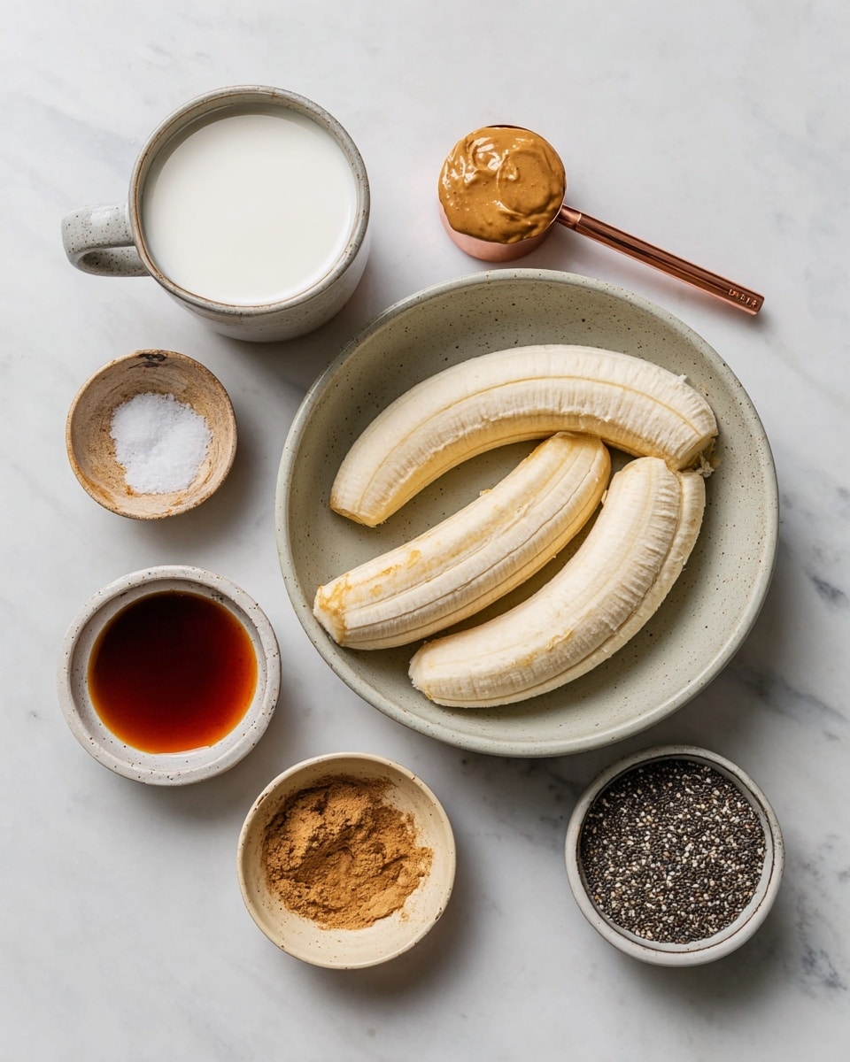 A top view of ingredients on a white marbled surface including four peeled frozen bananas placed inside a light gray bowl at the center, a ceramic cup filled with white milk on the upper left, a copper measuring spoon with a creamy light brown nut butter on the upper right, a small white bowl with ground cinnamon below it, a small round light beige bowl with coarse salt near the lower left, a similar light beige bowl with a dark brown liquid below it, and a small round bowl with black and white chia seeds near the lower right photo taken with an iphone --ar 4:5 --v 7