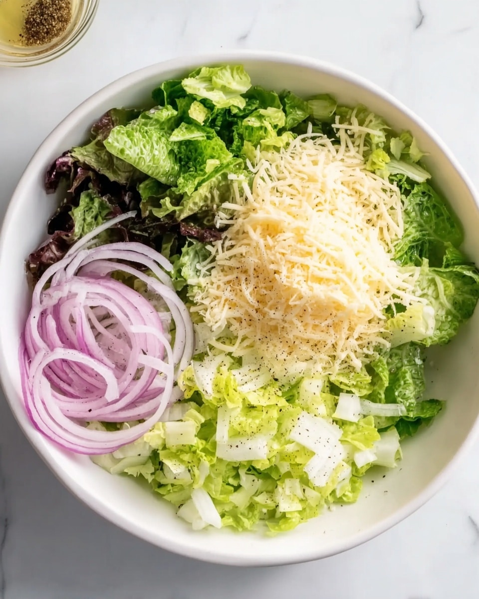 A white bowl on a white marbled surface holds layers of fresh ingredients arranged side by side. On the left side, there is a pile of dark green leafy lettuce with a crisp texture. Next to it on the top right, there is a mound of finely shredded pale yellow cheese. Below the cheese, on the bottom right side, there are light green chopped lettuce pieces. On the far left bottom corner inside the bowl, there are thin, curled slices of pale purple onion resting gently. A light dressing or oil with black pepper specks sits beneath the ingredients. Photo taken with an iphone --ar 4:5 --v 7