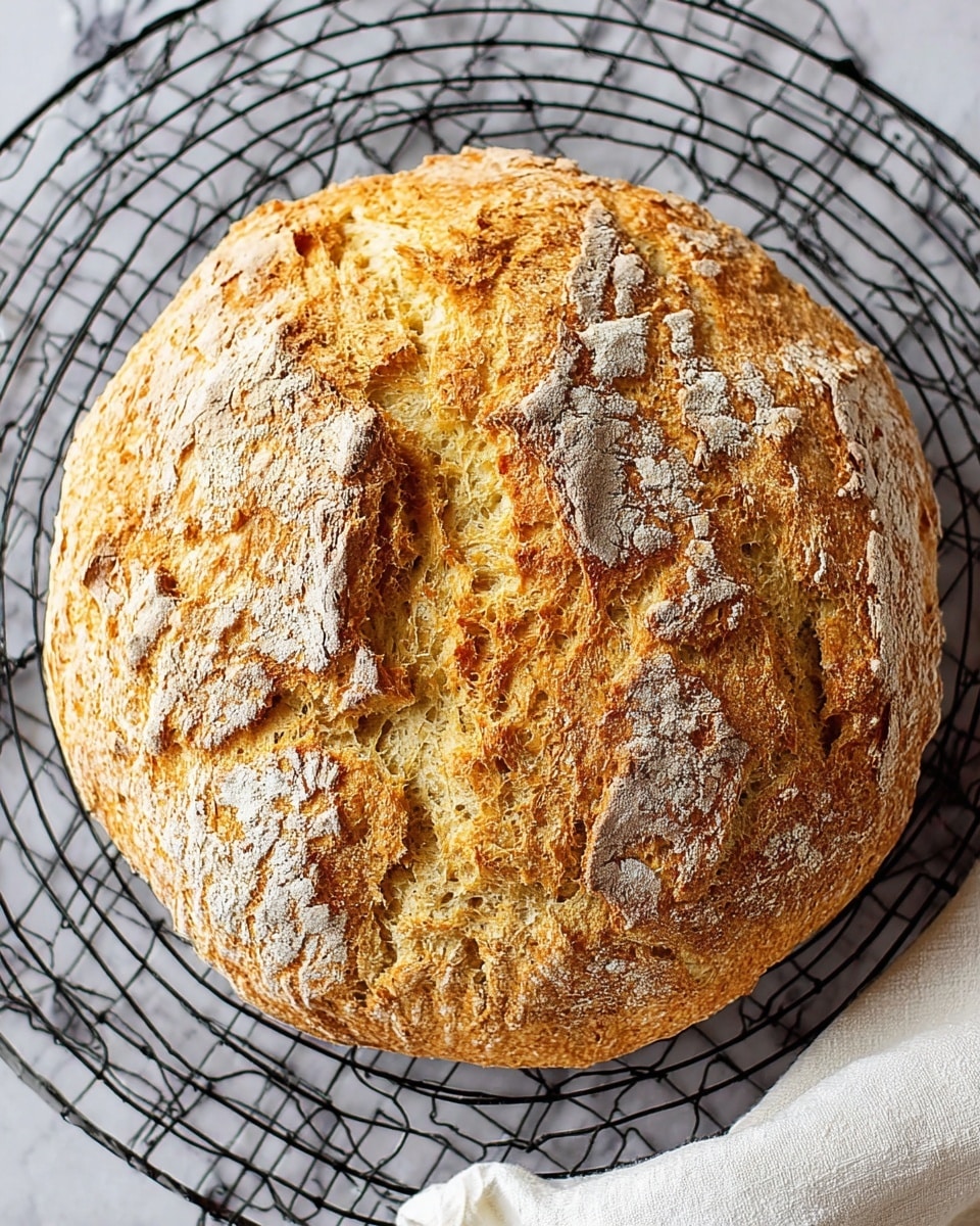 A round loaf of bread with a rough, golden-brown crust sits on a black cooling rack with a grid pattern, placed on a white marbled surface. The bread has a cracked texture with uneven crests, some patches dusted lightly with flour, and a split running down the center showing its soft, pale yellow inside. A folded white cloth is visible in the bottom right corner. Photo taken with an iphone --ar 4:5 --v 7