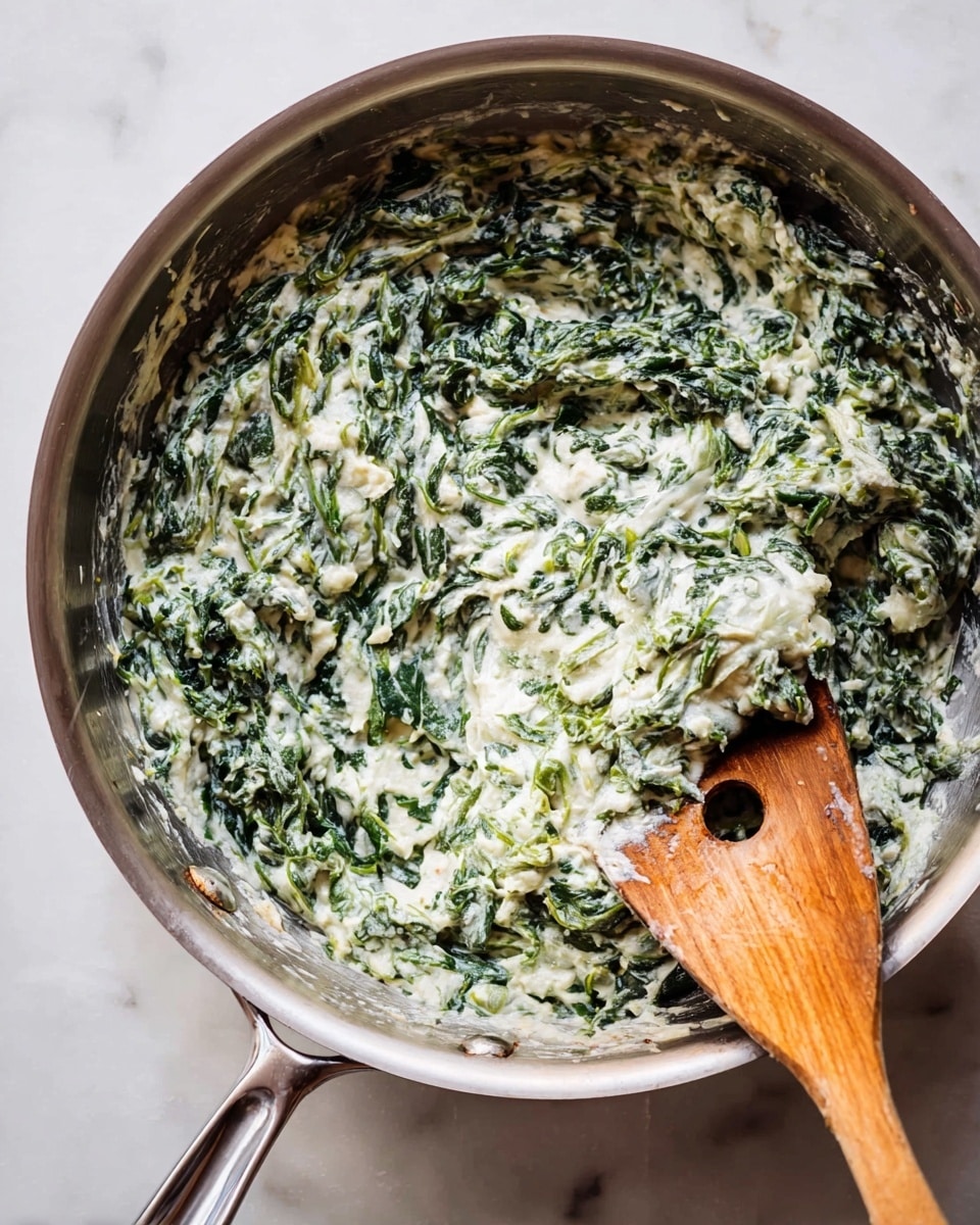 A close-up view of a stainless steel pan filled with a creamy spinach dish, showing a mix of dark green spinach leaves and soft white cream evenly blended together, creating a textured and slightly chunky surface; a wooden spoon with a hole in its center rests partially inside the pan on the right side, with some of the mixture clinging to its edge. The pan sits on a white marbled surface. photo taken with an iphone --ar 4:5 --v 7
