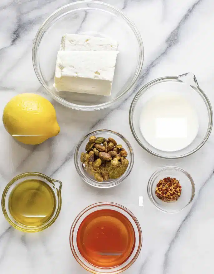 A clear food processor bowl filled with a base layer of yellow olive oil, topped with uneven chunks of white soft cheese scattered around the sharp central blade, all placed on a white marbled surface with a striped cloth slightly visible on the upper left side, photo taken with an iphone --ar 4:5 --v 7