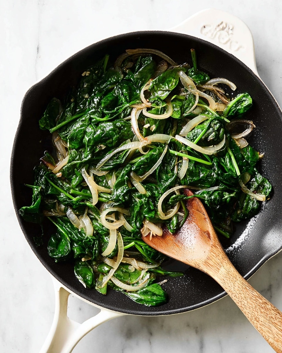 A black cooking pan with a white handle sits on a white marbled textured surface, filled with lightly cooked spinach and thinly sliced onions. The spinach leaves are deep green with a shiny, soft texture, scattered evenly across the pan. The thin onion slices are pale, slightly translucent with some light brown hints, mixed in among the spinach. A wooden spoon with a natural grain texture rests inside the pan, partially lifting some spinach and onions. The scene is bright and clean-looking, emphasizing the fresh and simple ingredients photo taken with an iphone --ar 4:5 --v 7