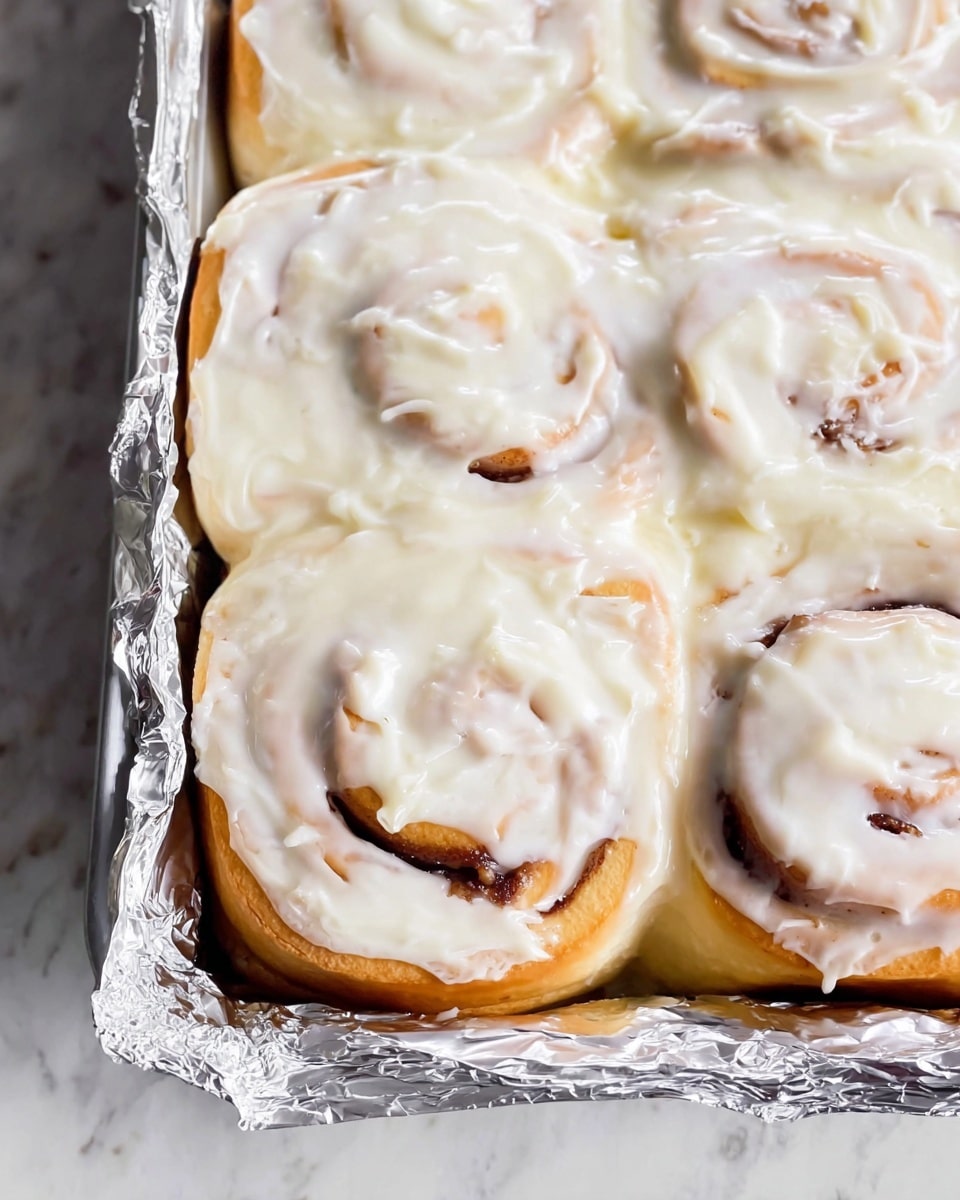 A close-up of a baking pan filled with several cinnamon rolls layered side by side, covered with a thick, smooth layer of white creamy icing that slightly melts and drips over the swirls of the rolls. The cinnamon rolls show a light golden-brown color underneath the creamy frosting with visible spirals of cinnamon filling. The pan is lined with crumpled shiny aluminum foil at the bottom, adding texture contrast to the soft and smooth icing and dough. The entire scene is set on a white marbled surface photo taken with an iphone --ar 4:5 --v 7