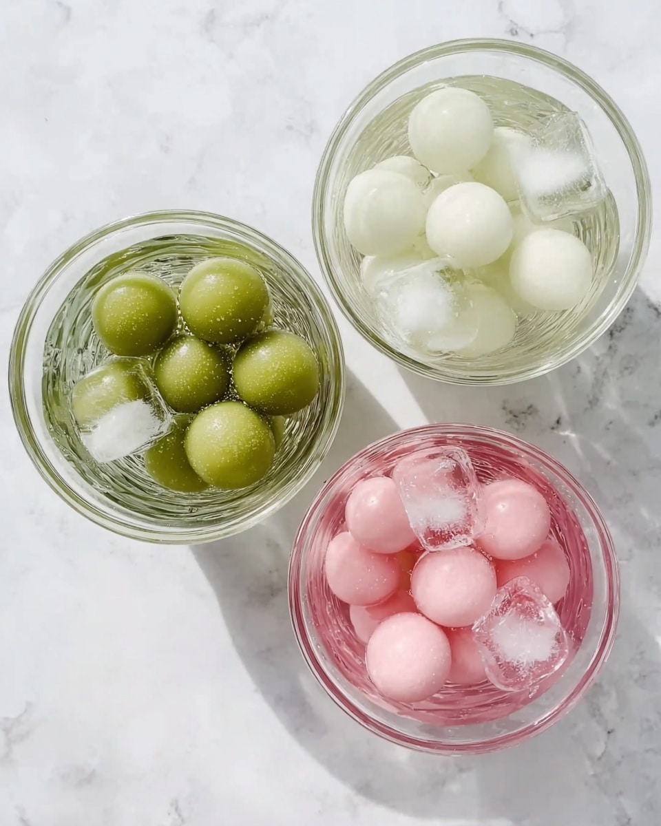 Three transparent glass bowls sit on a white marbled surface, each filled with water and ice cubes. The top left bowl holds several green round balls with a smooth texture, floating in the water with pieces of ice. The top right bowl contains pink round balls, also smooth, immersed in water with ice cubes. The bottom center bowl shows white round balls, similar in size and texture to the others, floating with ice cubes in water. The setup is clean and neatly arranged, viewed from above. photo taken with an iphone --ar 4:5 --v 7