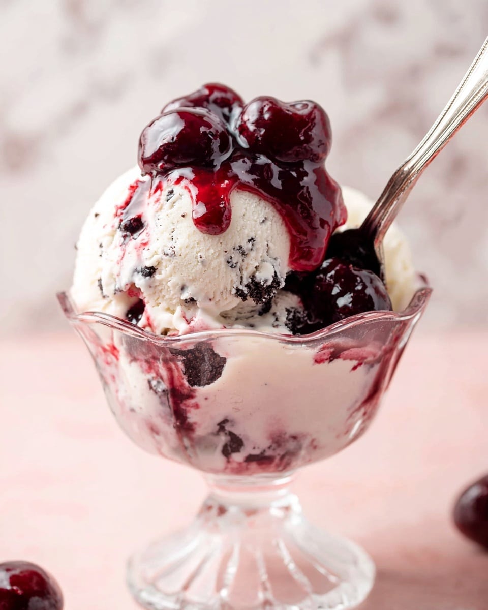 The image shows a clear glass dessert cup filled with one large scoop of white ice cream with dark cookie bits mixed inside, creating a marbled texture. On top, there is a thick layer of dark red cherry sauce with shiny whole cherries, some juice dripping slightly over the ice cream edges. A silver spoon is partly stuck into the right side of the ice cream, visible just above the surface. The dessert cup is placed on a soft pink surface with a blurred white marbled texture background. photo taken with an iphone --ar 4:5 --v 7