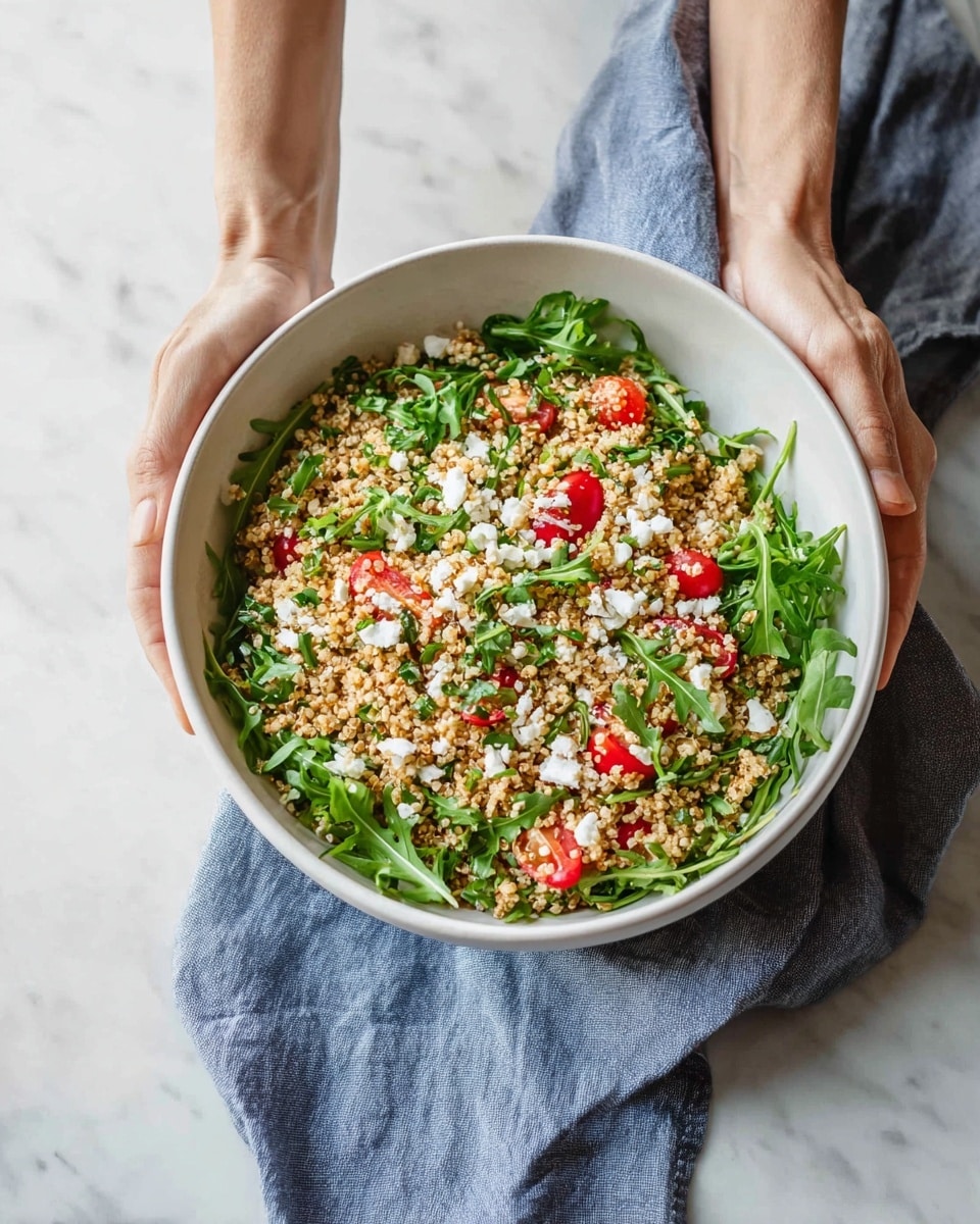 A white bowl filled with a three-layer salad is held by a woman's hands over a blue-grey cloth on a white marbled surface. The bottom layer is fresh green arugula leaves spread evenly across the bowl. The middle layer has scattered bright red cherry tomato halves peeking through. The top layer consists of a large amount of light brown cooked quinoa speckled with small white crumbles of cheese, giving a textured look. The salad looks fresh and colorful with a mix of smooth tomatoes, grainy quinoa, and leafy greens. Photo taken with an iphone --ar 4:5 --v 7
