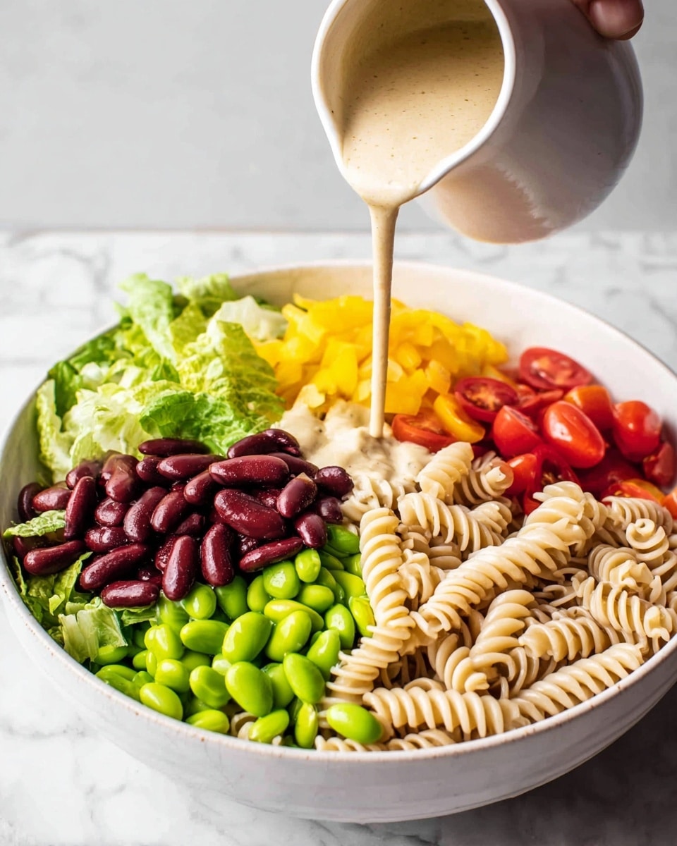 A white bowl sits on a white marbled surface and holds a colorful layered salad with seven distinct sections: leafy green lettuce on the top left, bright green edamame beans just below the lettuce, dark red kidney beans next to the edamame, pale beige spiral pasta on the bottom right, yellow diced bell peppers above the pasta, and small halved red cherry tomatoes next to the bell peppers. A creamy beige dressing is being poured over the salad from a white pitcher, creating a smooth layer over the edamame and some nearby ingredients. Woman's hand holds the pitcher. Photo taken with an iphone --ar 4:5 --v 7