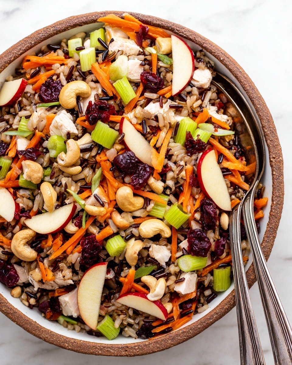 A bowl filled with a colorful mixed salad showing three main layers: the bottom layer with brown and wild rice, the middle layer with small chopped cubes of white chicken, light green celery pieces, and small chunks of red apple, and the top layer sprinkled with bright orange shredded carrot, white cashew nuts, and dark red dried cranberries. The bowl is white with a slightly visible ornate pattern on its outer side, placed on a white marbled surface with a glass jar of creamy dressing visible at the upper left corner. photo taken with an iphone --ar 4:5 --v 7