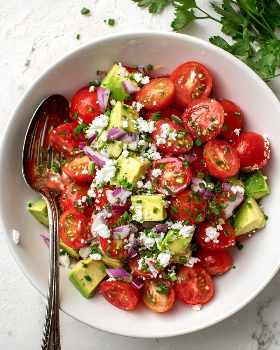 A white bowl filled with a fresh salad showing three main layers: bright red cherry tomato halves on top, sprinkled with small green chives; cubes of light green avocado pieces mixed evenly in the middle; and white crumbled cheese scattered generously over the salad; small pieces of finely chopped purple onion spread throughout; a vintage silver slotted spoon resting inside the bowl on the left side; background and surface are white marbled texture with some green leafy herbs on the top right corner photo taken with an iphone --ar 4:5 --v 7